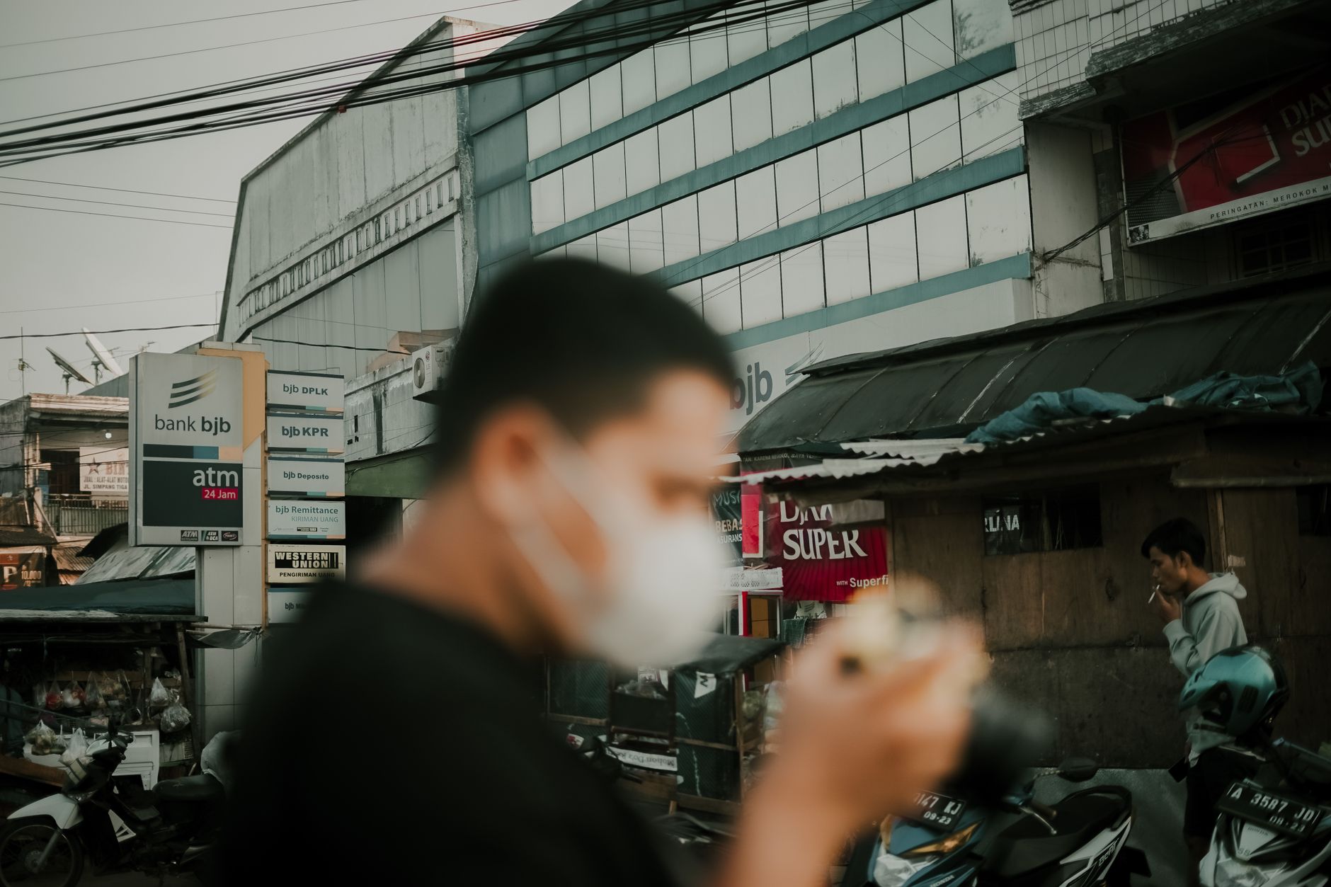 A person in a face mask walks down a busy city street past a bank bjb ATM sign.