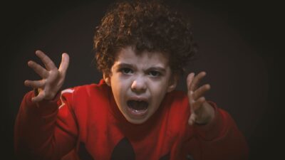 A young boy with curly hair in a red sweater screaming in frustration, representing a child tantrum.