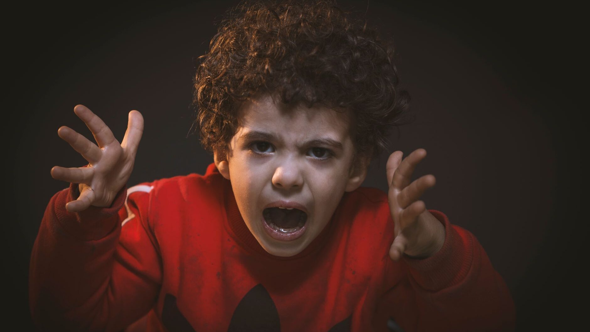 A young boy with curly hair in a red sweater screaming in frustration, representing a child tantrum.