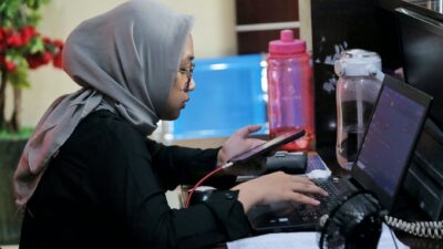 A woman in a grey hijab focused on her laptop, demonstrating tech adaptation at her desk.