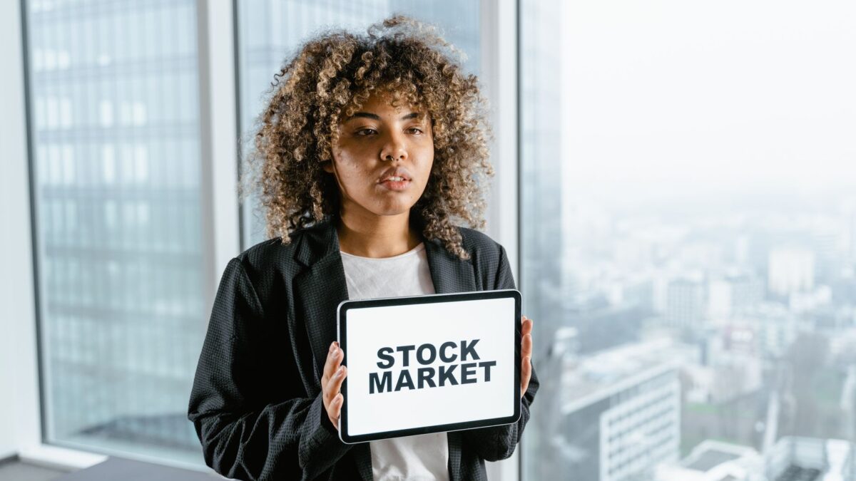 A young woman with curly hair holds a tablet showing "STOCK MARKET" in an office, illustrating Mitos Investasi Saham.