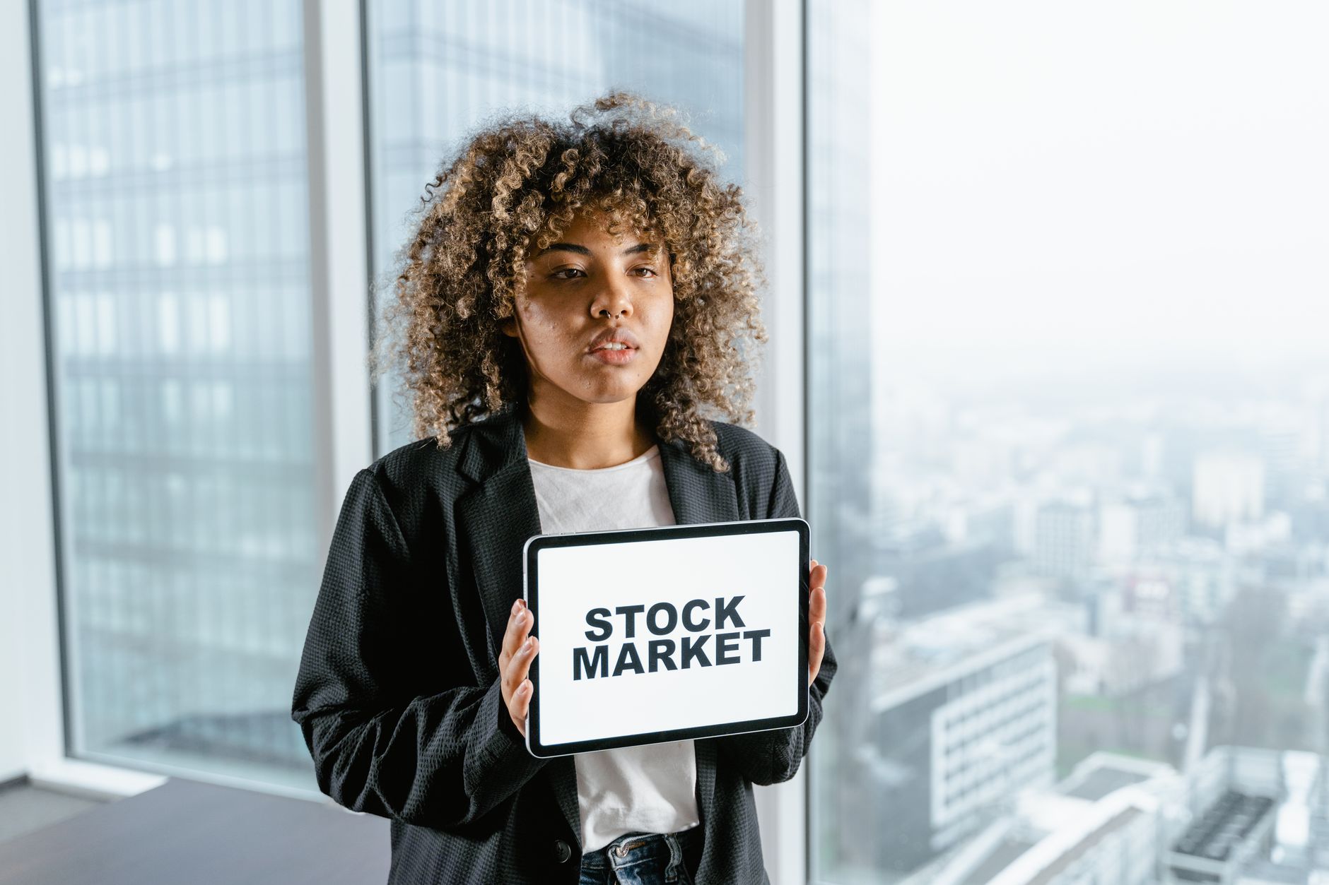 A young woman with curly hair holds a tablet showing "STOCK MARKET" in an office, illustrating Mitos Investasi Saham.