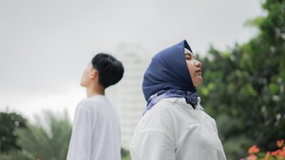 A woman in a blue hijab and a man stand outdoors facing opposite directions, highlighting the Perbedaan Belahan Jiwa.