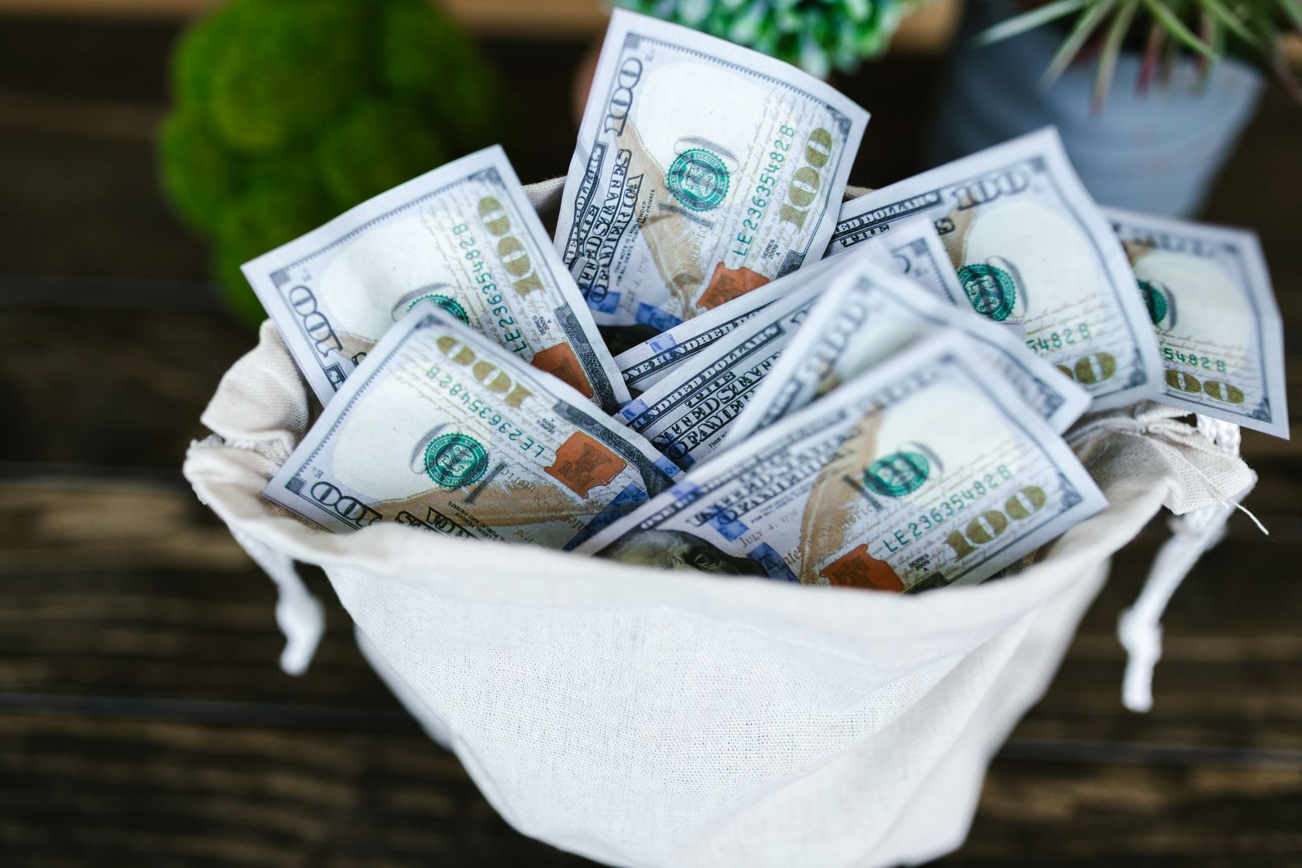 A drawstring pouch filled with US hundred dollar bills on a wooden table.