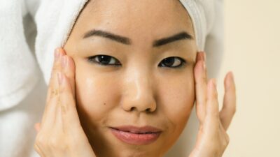 Wanita Asia tersenyum sambil melakukan perawatan wajah dengan handuk di kepala.