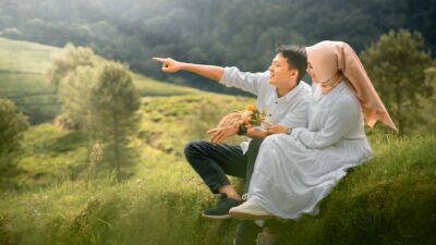 A couple in white sits on a grassy hill overlooking a valley, illustrating the complexities of pencarian pasangan.