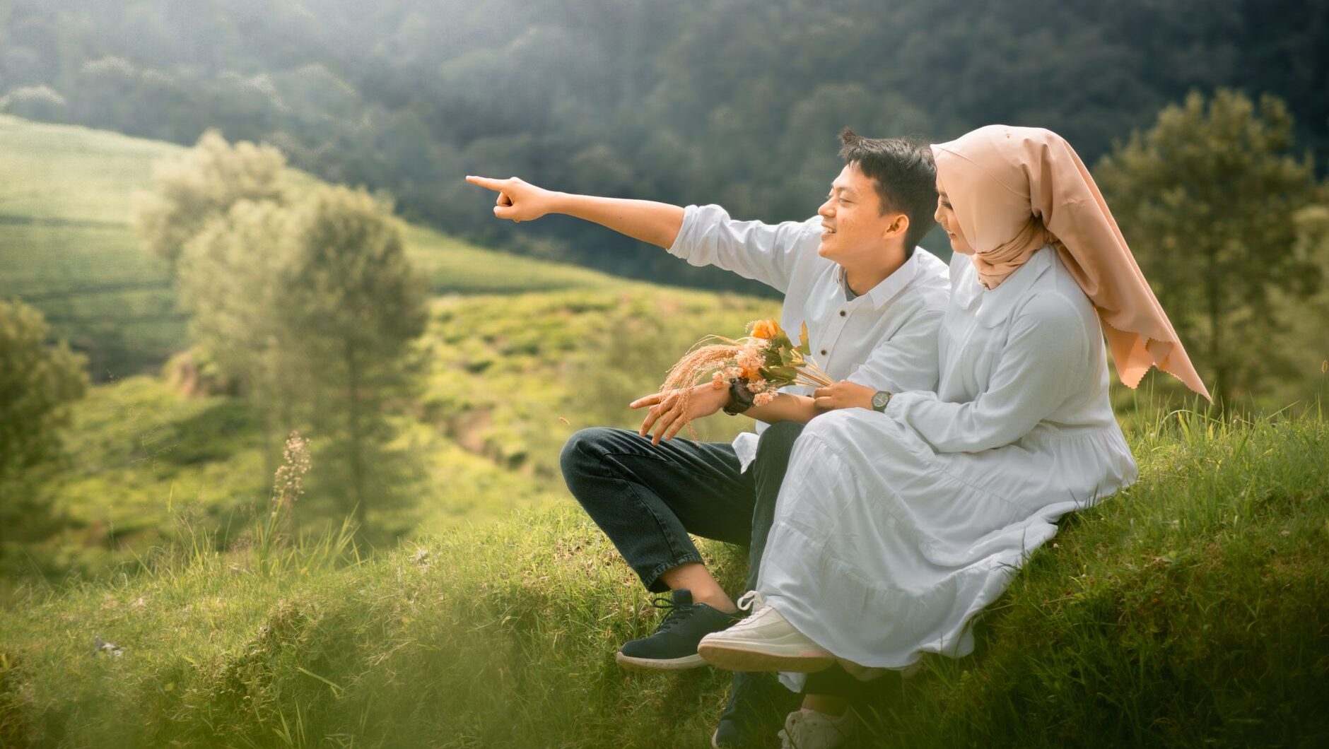 A couple in white sits on a grassy hill overlooking a valley, illustrating the complexities of pencarian pasangan.
