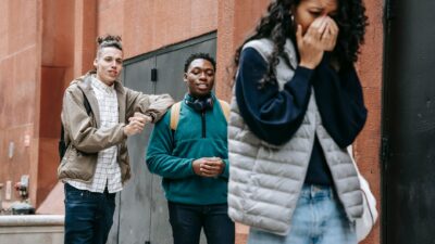 Two boys laughing and pointing at a distressed teenage girl covering her face, depicting peer bullying.