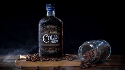 Botol kopi "Cold Brew" dikelilingi biji kopi sangrai di papan kayu, dengan gelas terguling yang tumpah di samping.