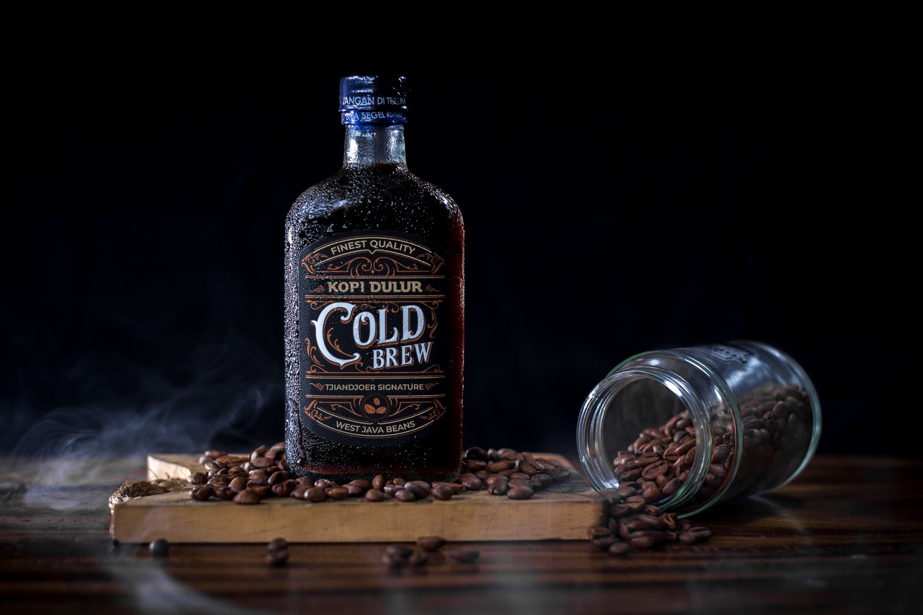Botol kopi "Cold Brew" dikelilingi biji kopi sangrai di papan kayu, dengan gelas terguling yang tumpah di samping.