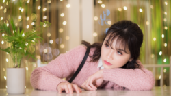 A pensive young Asian woman in a pink sweater rests her head on her arms at a table.