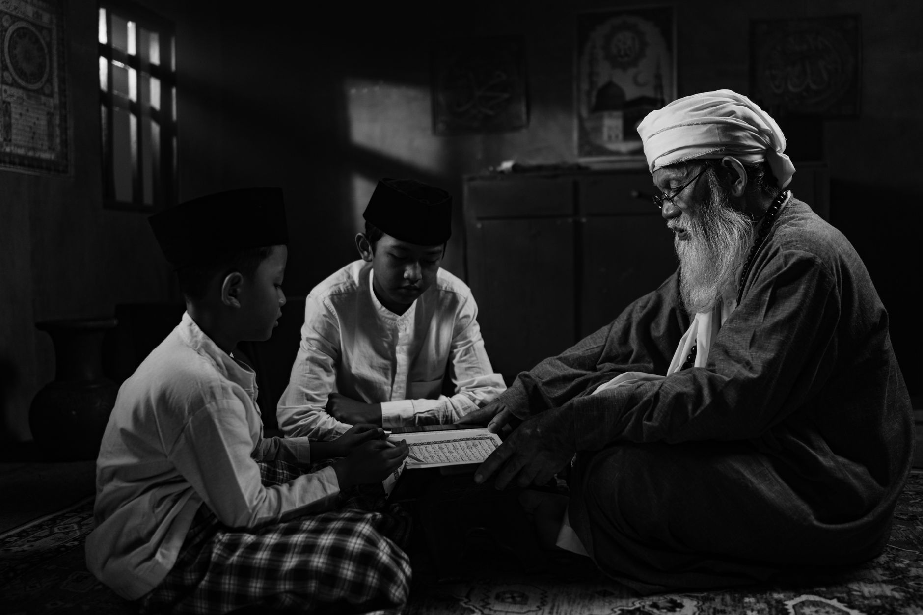 Elderly man guiding two young boys reading a holy book indoors, representing traditional nasihat orang tua.