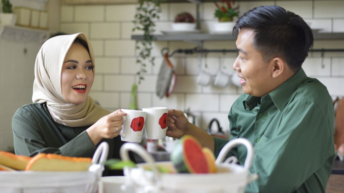 Muslim couple smiling and cheering with coffee cups in a cozy kitchen, representing Tanda Pasangan Jodoh.