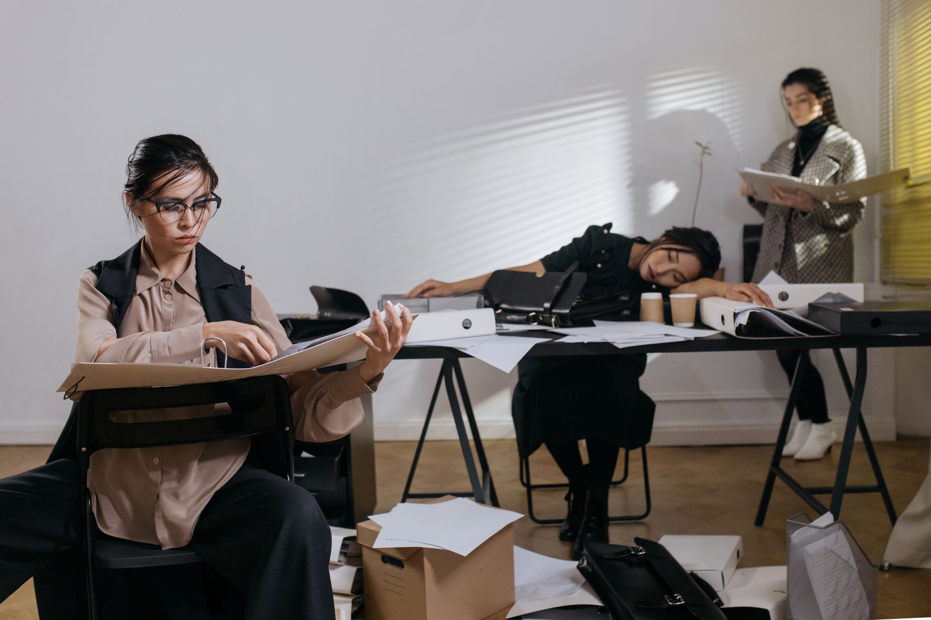 Three women in a messy office, with one woman sleeping at her desk while the others work.