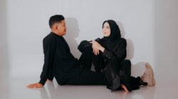 A young couple in black outfits smiles and looks at each other against a white wall.
