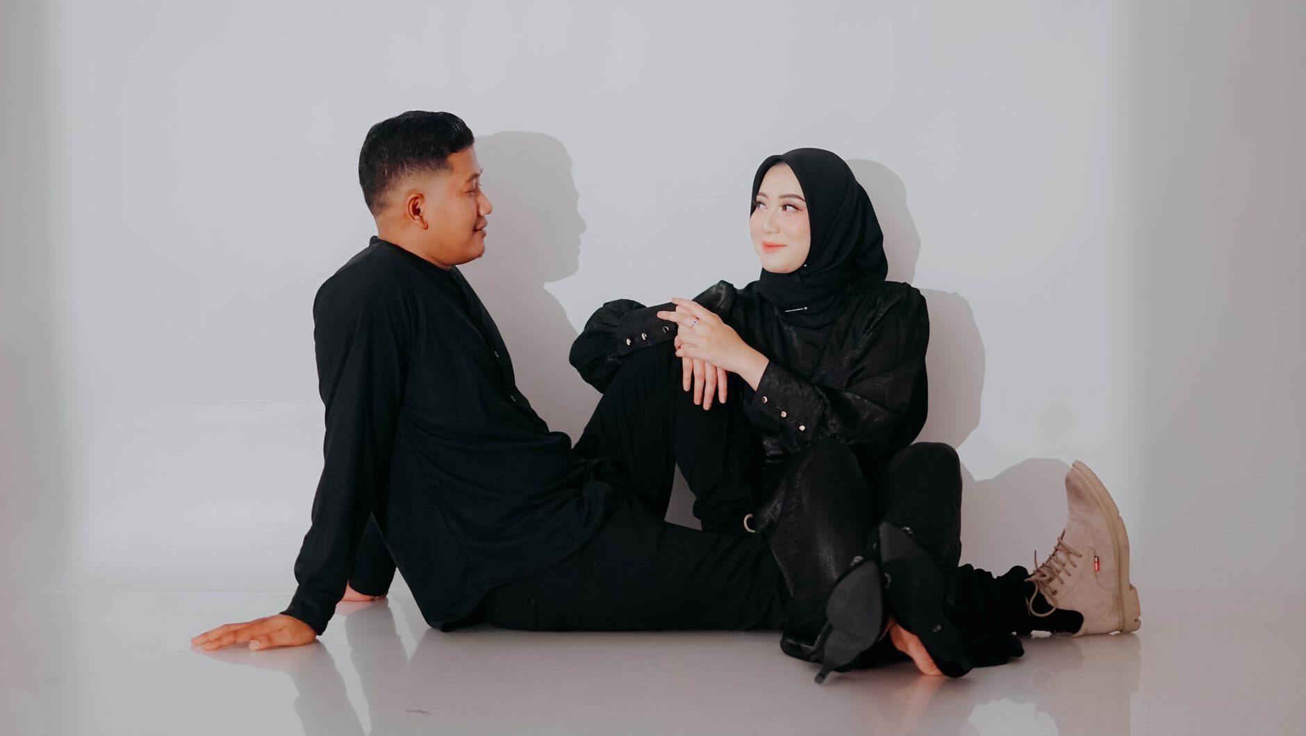 A young couple in black outfits smiles and looks at each other against a white wall.