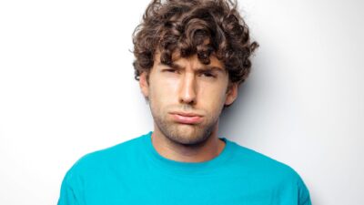 A young man with curly hair wearing a blue t-shirt makes a disappointed expression representing Irritable Male Syndrome.
