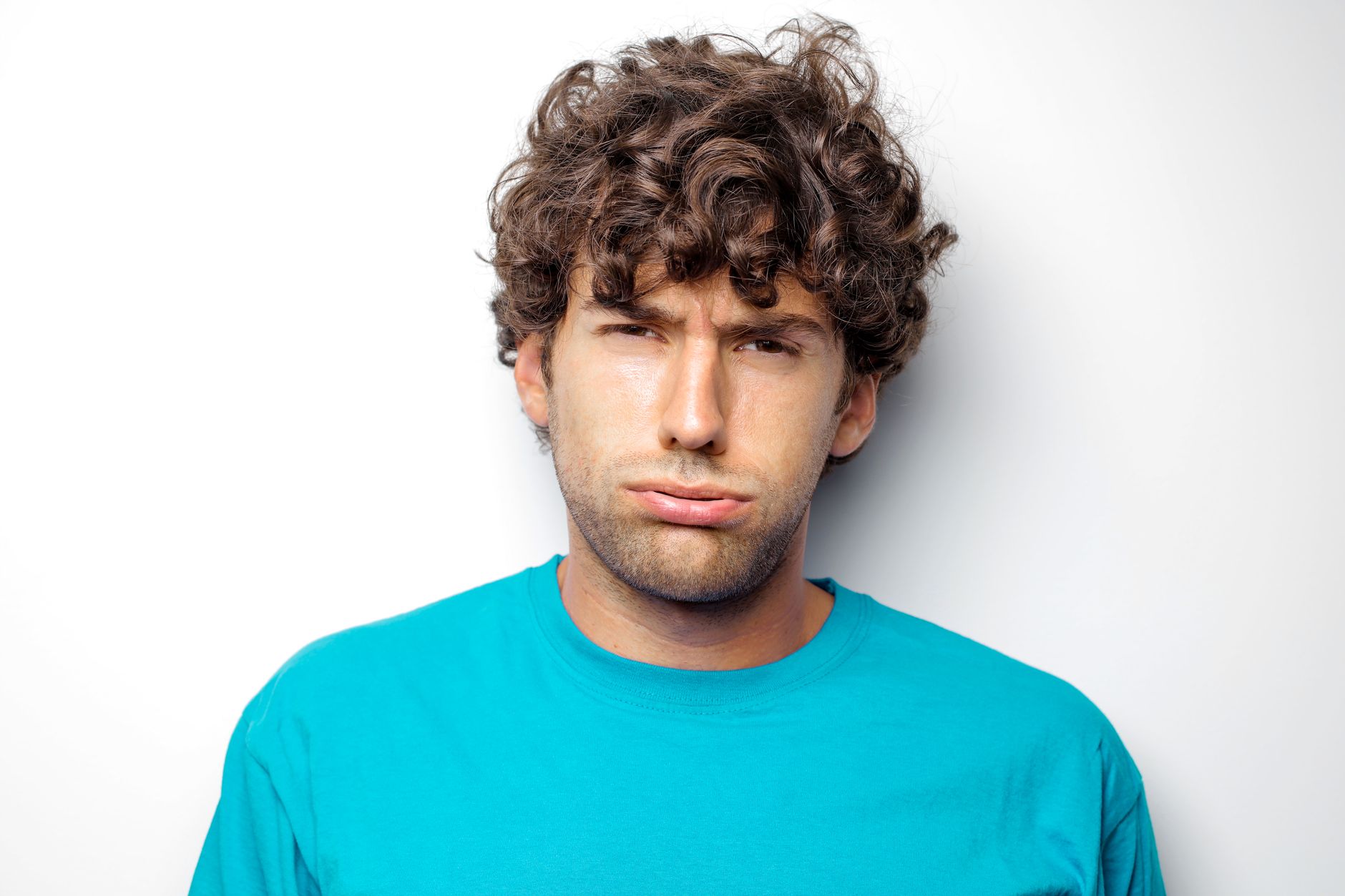 A young man with curly hair wearing a blue t-shirt makes a disappointed expression representing Irritable Male Syndrome.