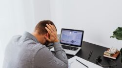 Seorang pria tampak stres di depan laptop dengan grafik keuangan, menggambarkan penyebab kesulitan finansial.