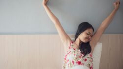Woman with dark hair stretching her arms overhead while waking up in a bed with white linens and a wooden headboard.