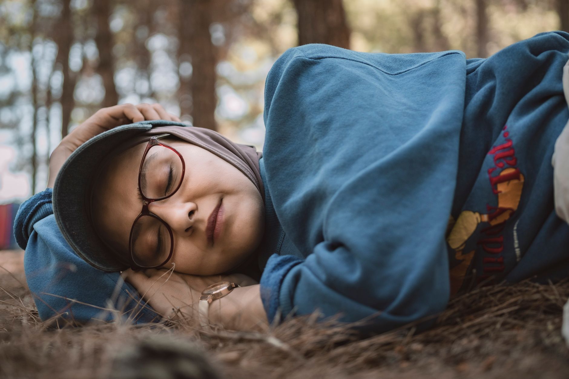 Wanita berhijab dan berkacamata tidur tenang di lantai hutan sebagai bagian dari pola istirahat.
