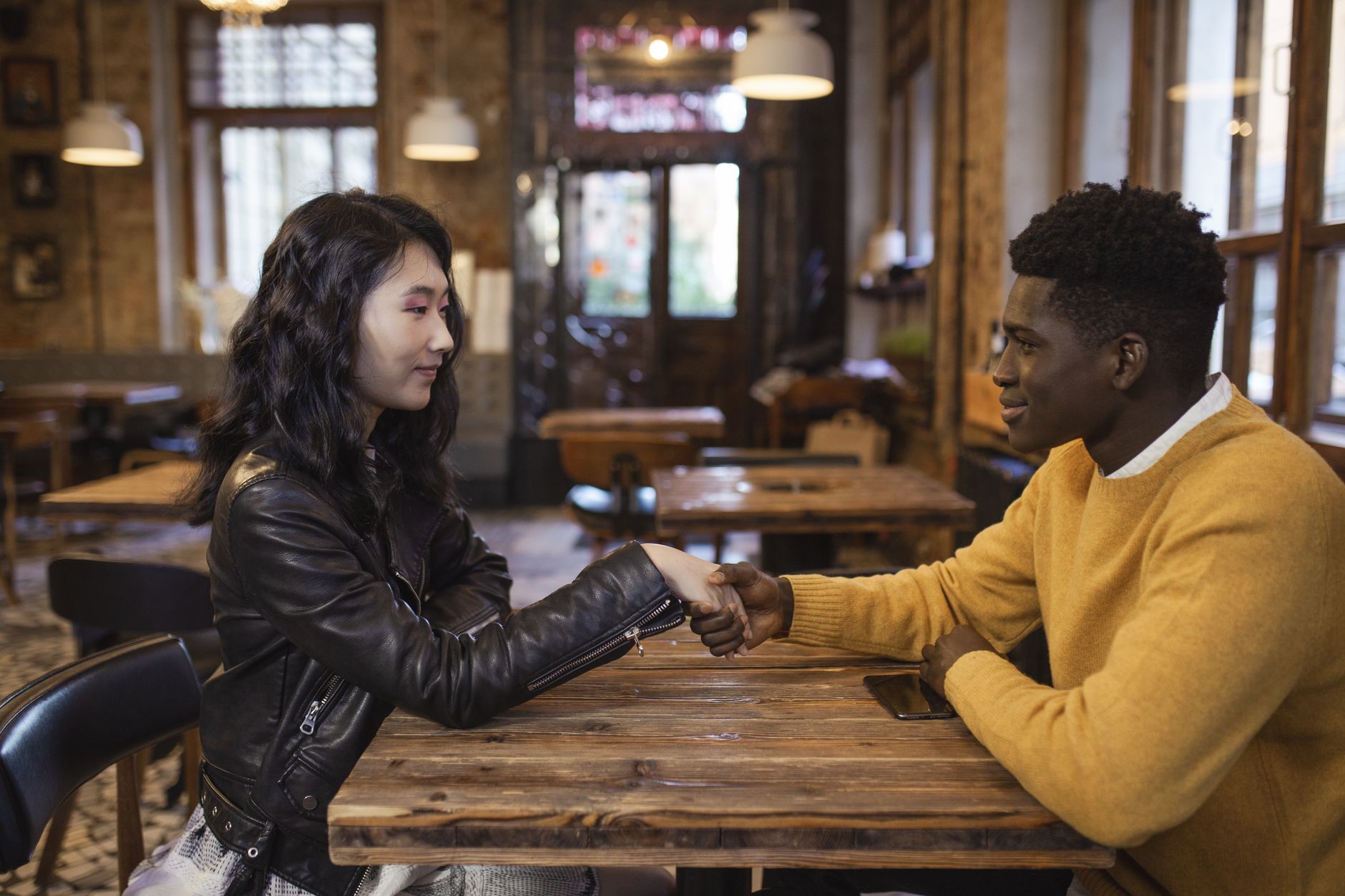 A young multi-ethnic couple holding hands across a wooden table in a cozy cafe, illustrating psikologi flirting.