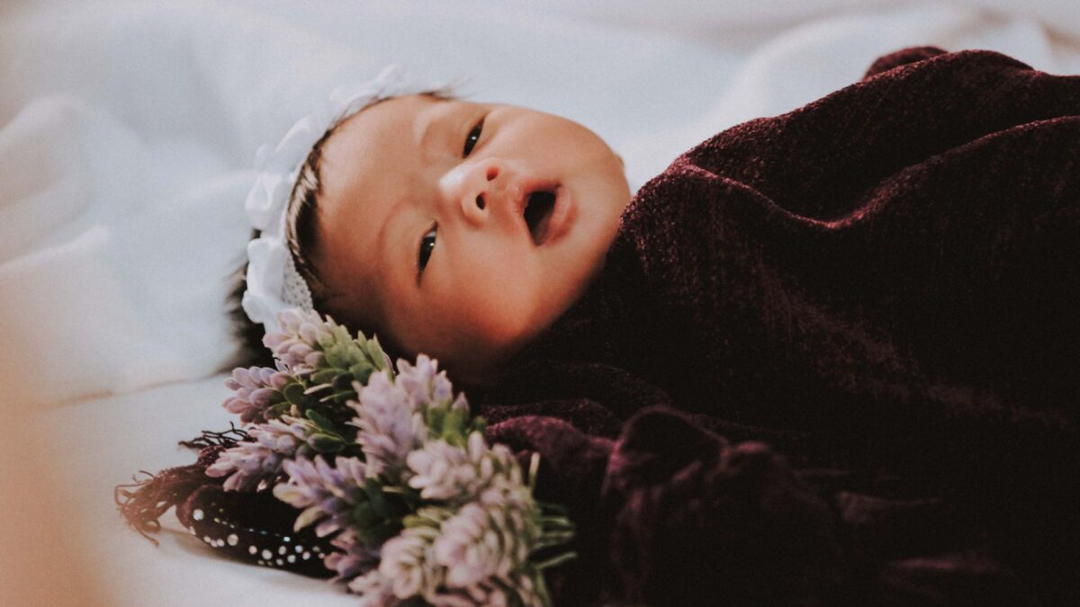 Bayi perempuan baru lahir tidur nyenyak dibalut selimut ungu di samping bunga.