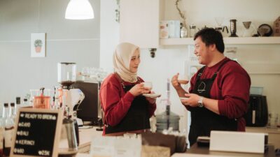Seorang wanita berjilbab krem dan atasan merah serta pria berapron gelap mengobrol sambil makan camilan di meja kafe.