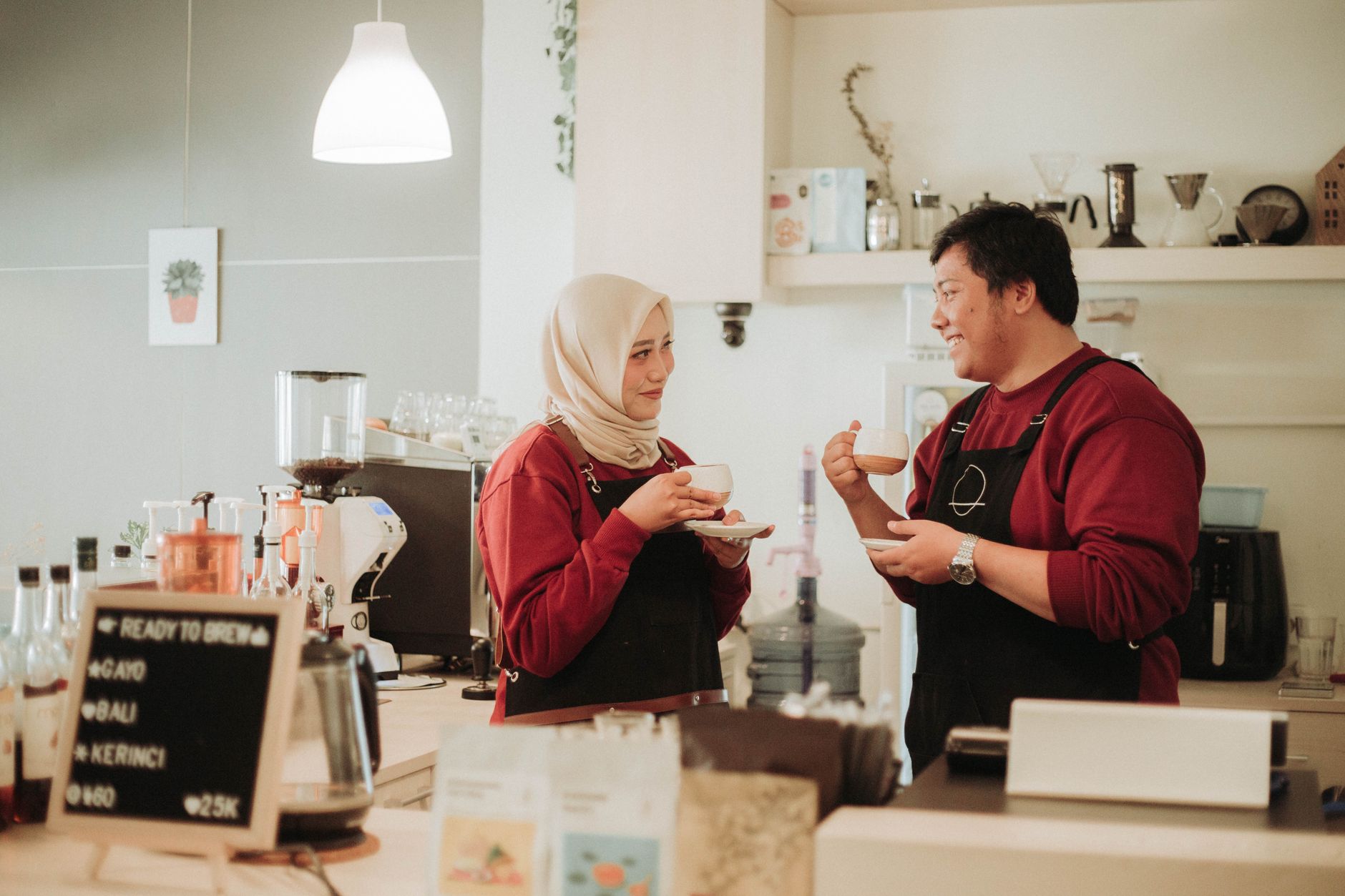 Seorang wanita berjilbab krem dan atasan merah serta pria berapron gelap mengobrol sambil makan camilan di meja kafe.
