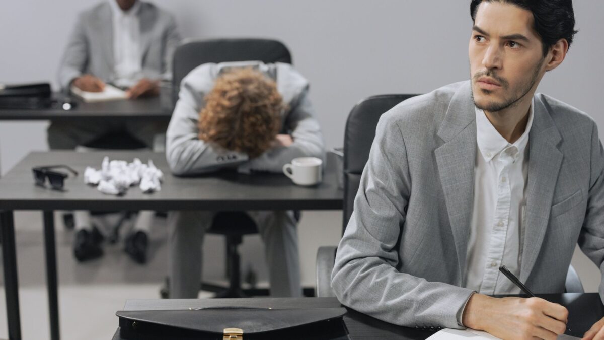 Businessman sleeping at his desk with crumpled paper while coworkers work in the background, illustrating Sindrom Impostor Ka