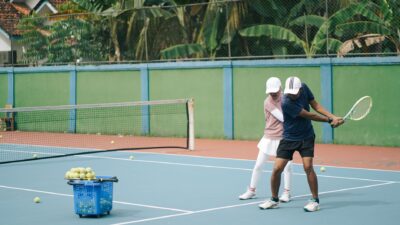 Seorang pelatih tenis membimbing pemain di lapangan luar ruangan yang biru dengan keranjang bola.