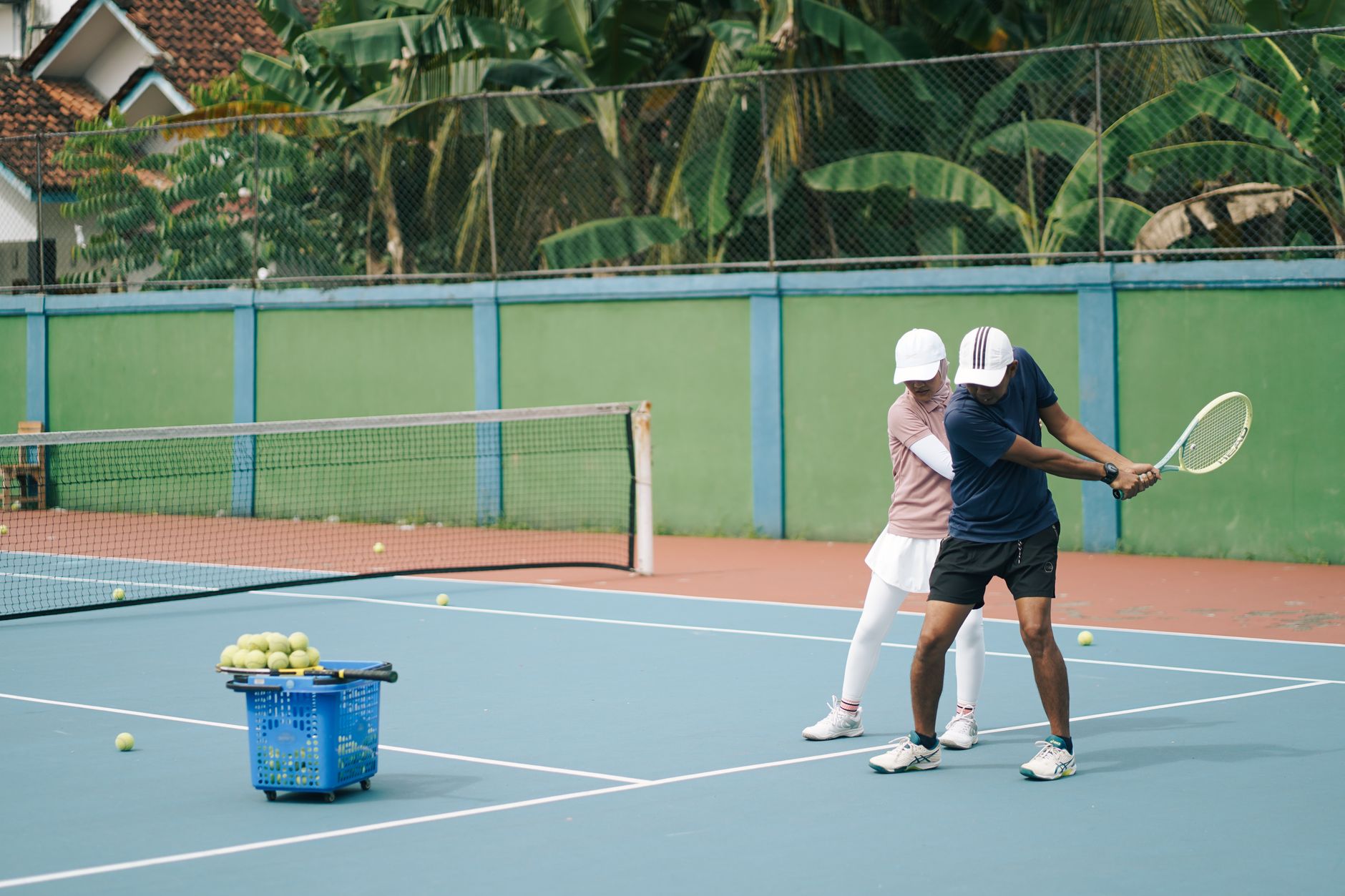 Seorang pelatih tenis membimbing pemain di lapangan luar ruangan yang biru dengan keranjang bola.