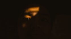 Close-up of a person's face in darkness, dramatically illuminated by a beam of light on one eye, representing Sisi Gelap Menj