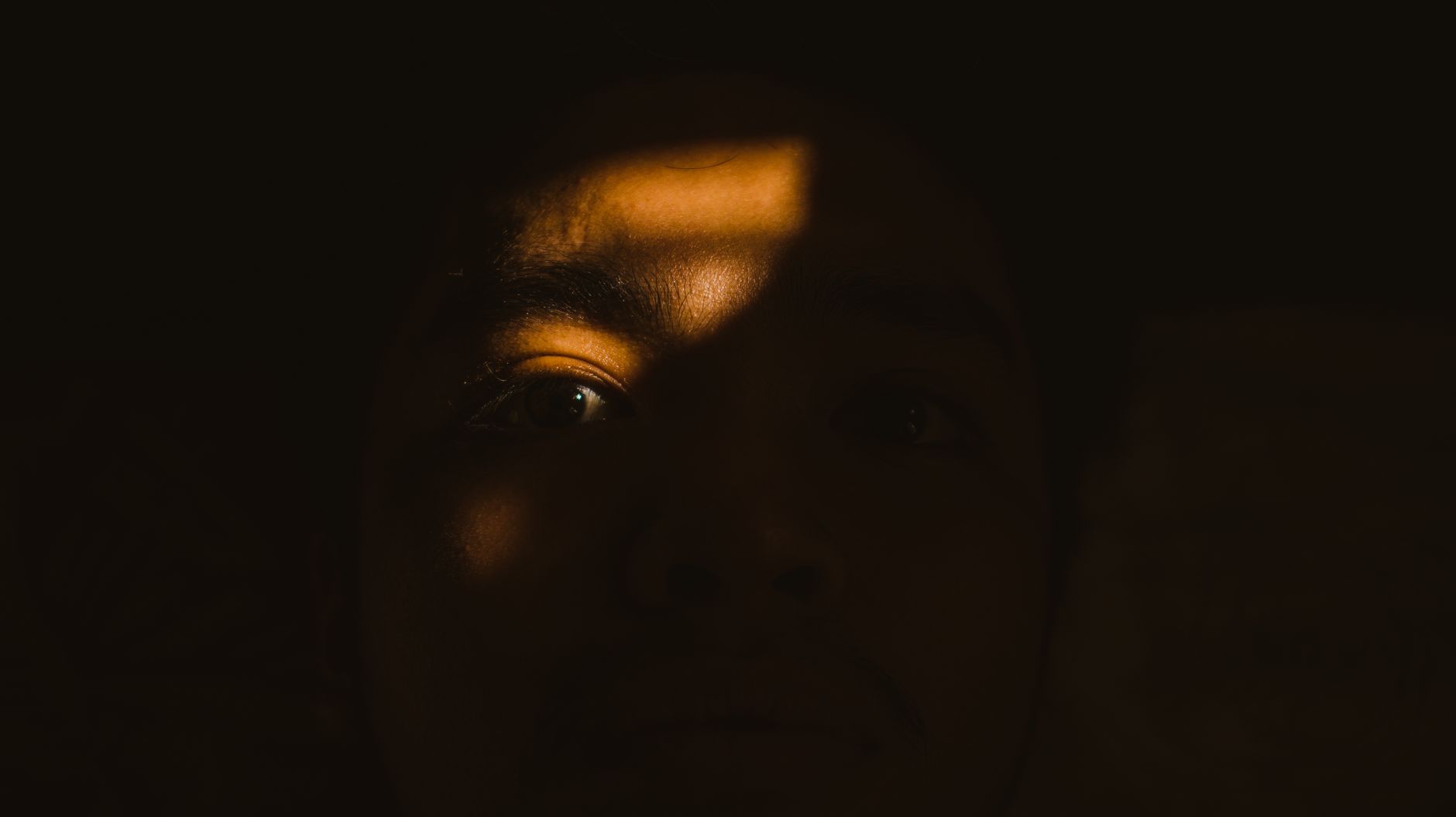 Close-up of a person's face in darkness, dramatically illuminated by a beam of light on one eye, representing Sisi Gelap Menj