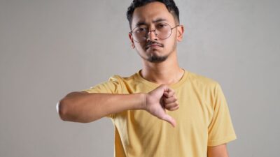 A South Asian man in a yellow t-shirt and glasses gives a thumbs-down gesture, showing disapproval.