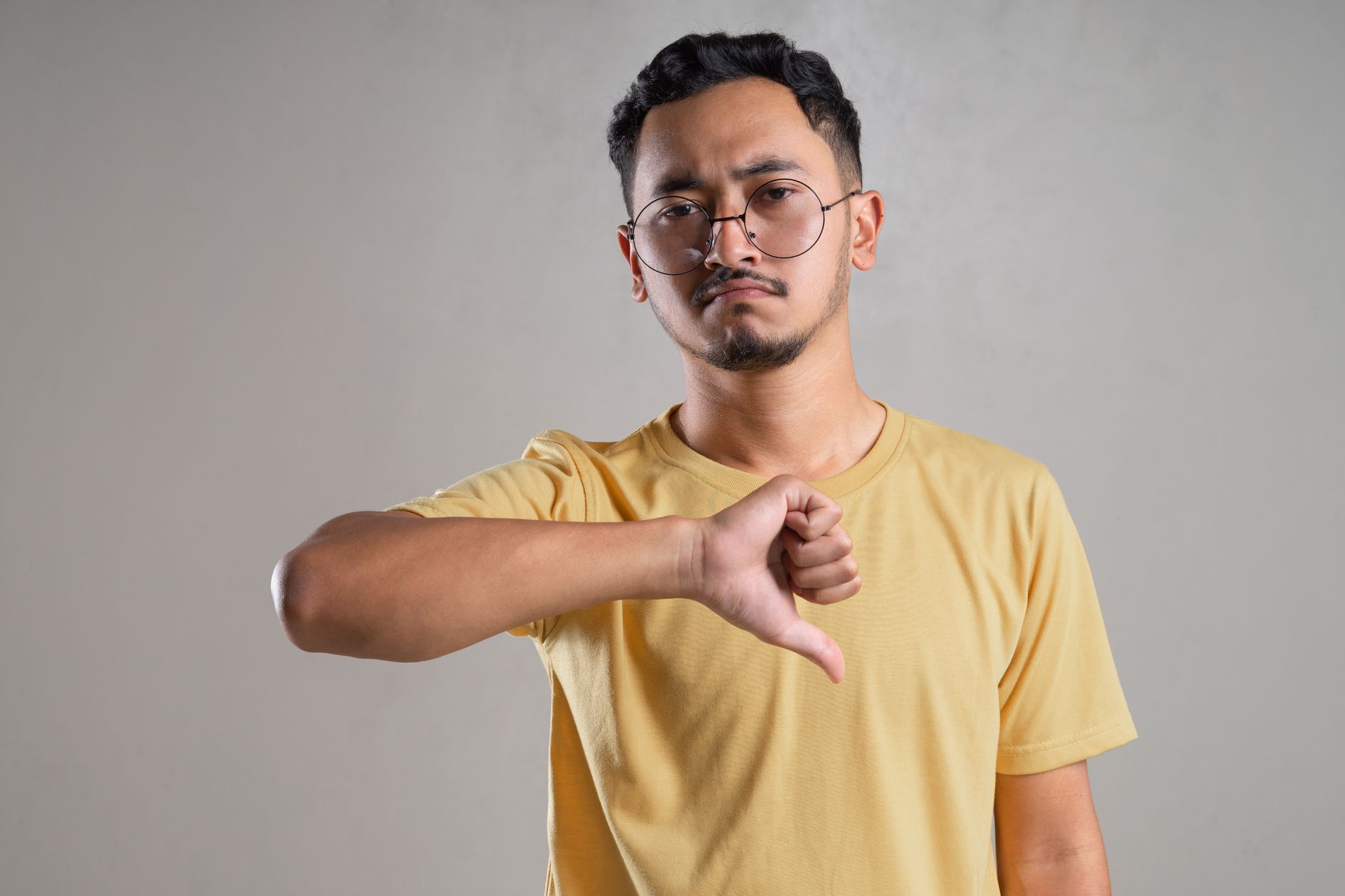 A South Asian man in a yellow t-shirt and glasses gives a thumbs-down gesture, showing disapproval.