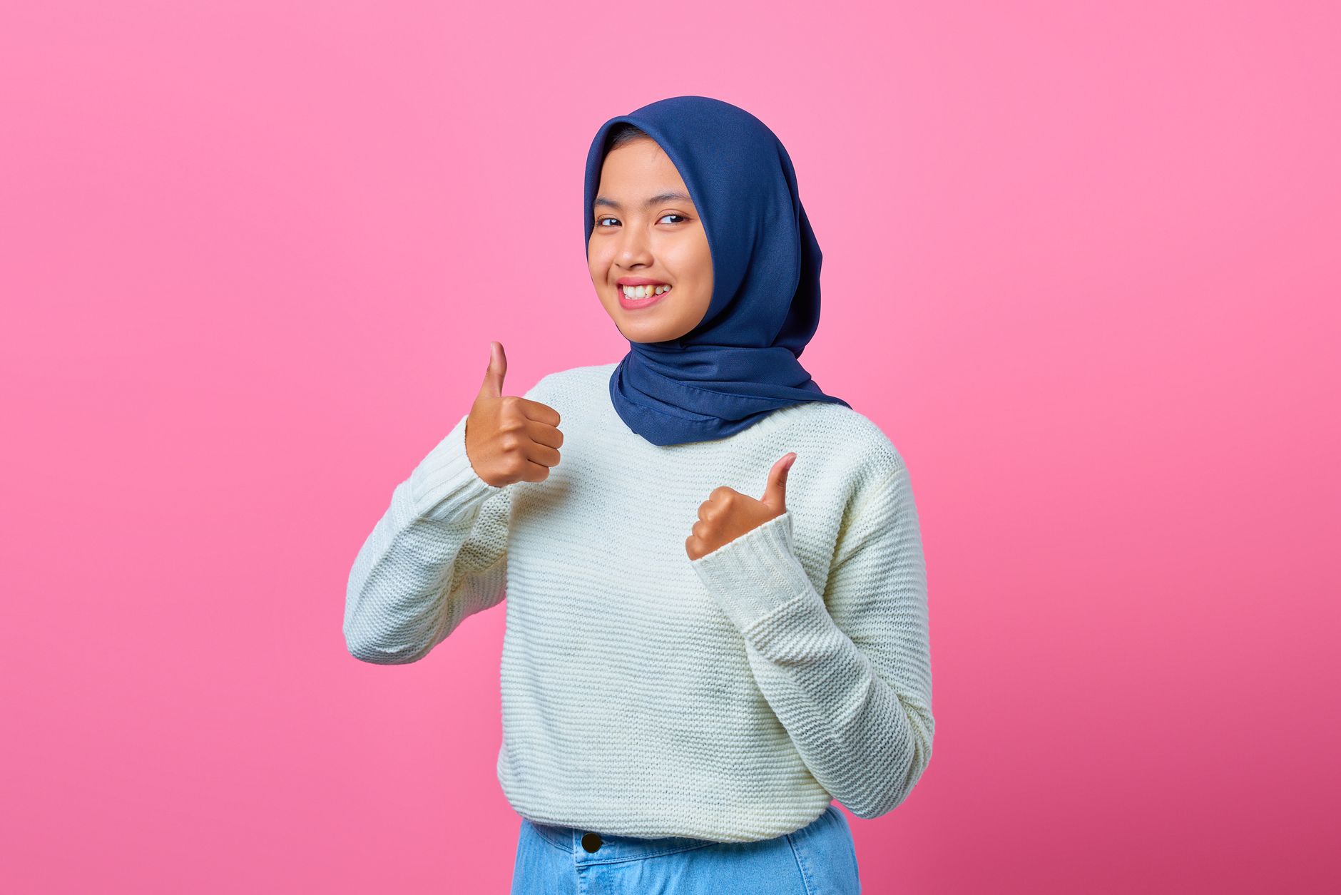 Smiling young Asian woman in blue hijab and white sweater giving two thumbs up.