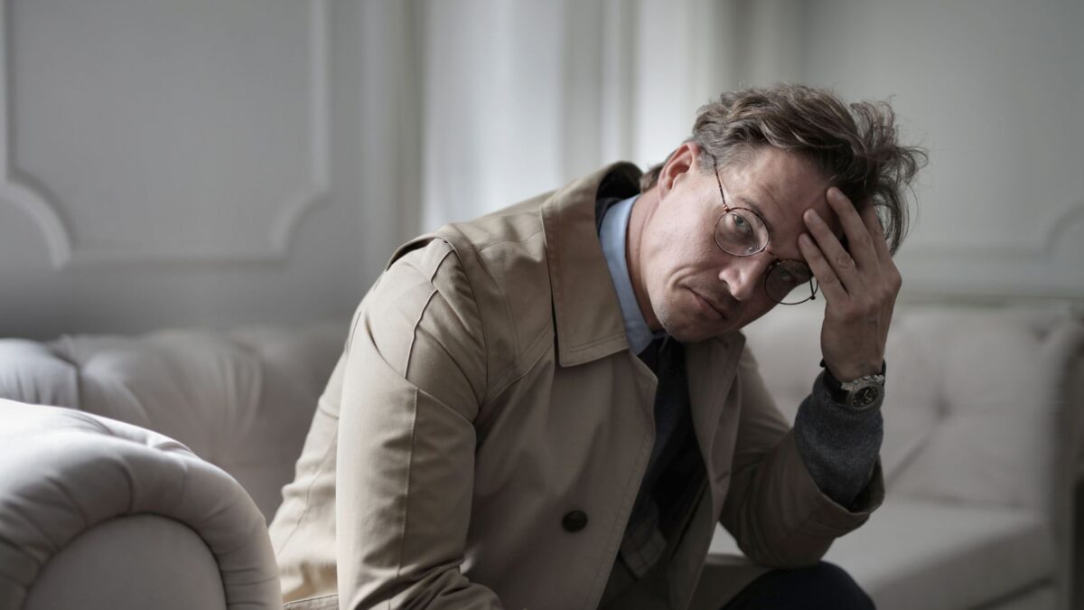 A stressed man in a trench coat holding his head while sitting on a couch, illustrating anxiety pensiun.