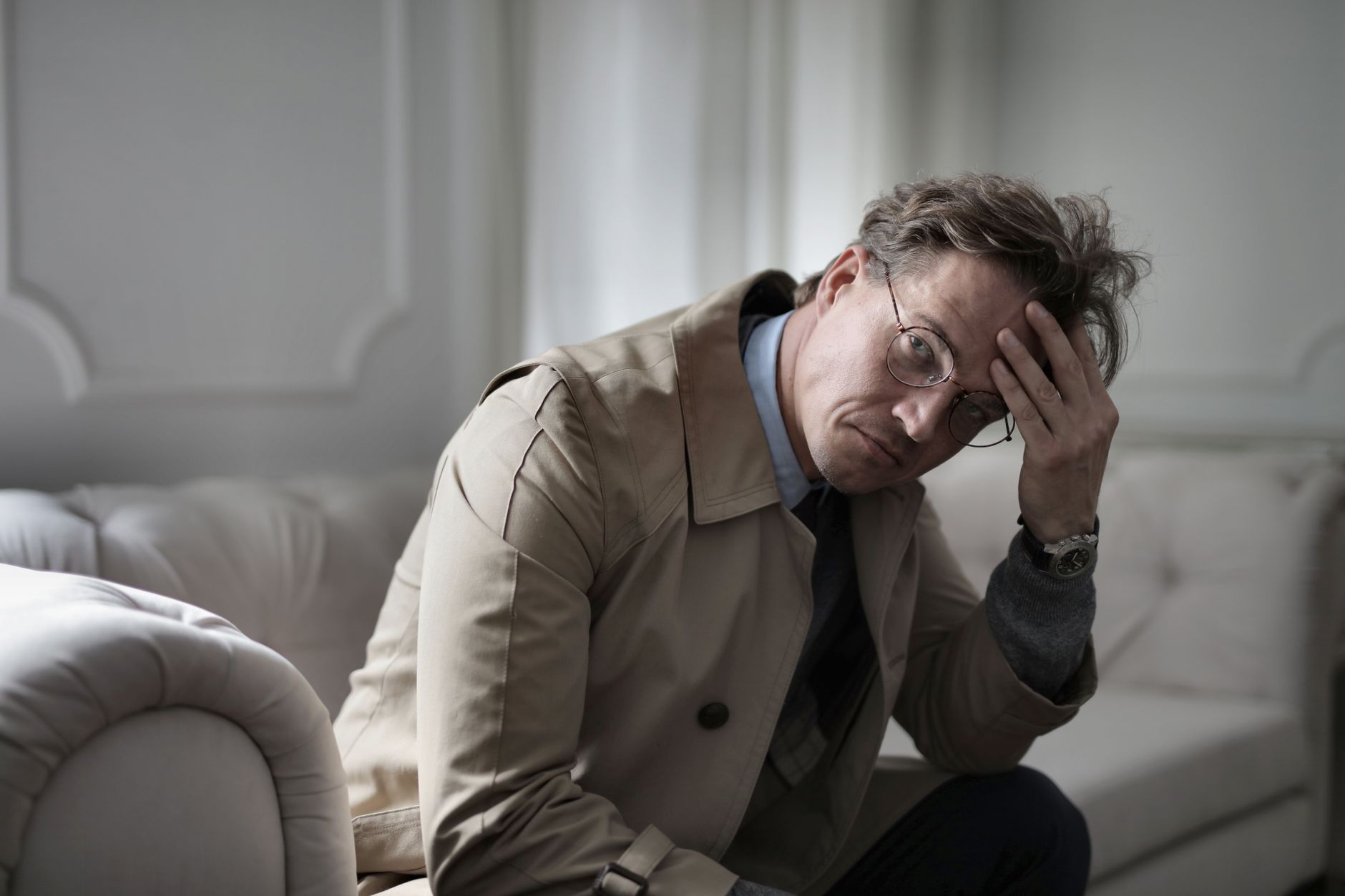A stressed man in a trench coat holding his head while sitting on a couch, illustrating anxiety pensiun.