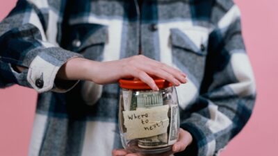 A person holding a glass jar labeled "Where to move?" filled with cash, illustrating frugal living strategies.
