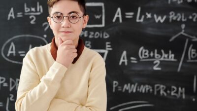 A thoughtful student wearing glasses stands in front of a chalkboard filled with math equations.