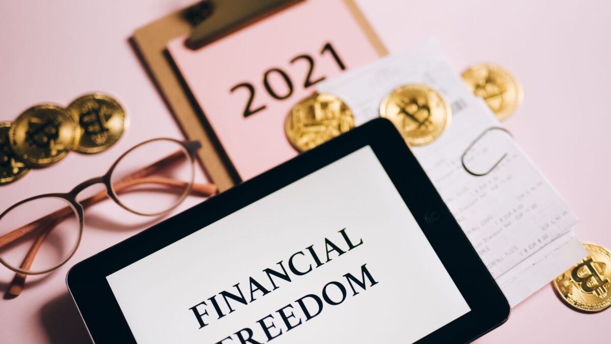 A tablet showing "Financial Freedom", a notebook, glasses, and gold bitcoin coins on a pink surface.