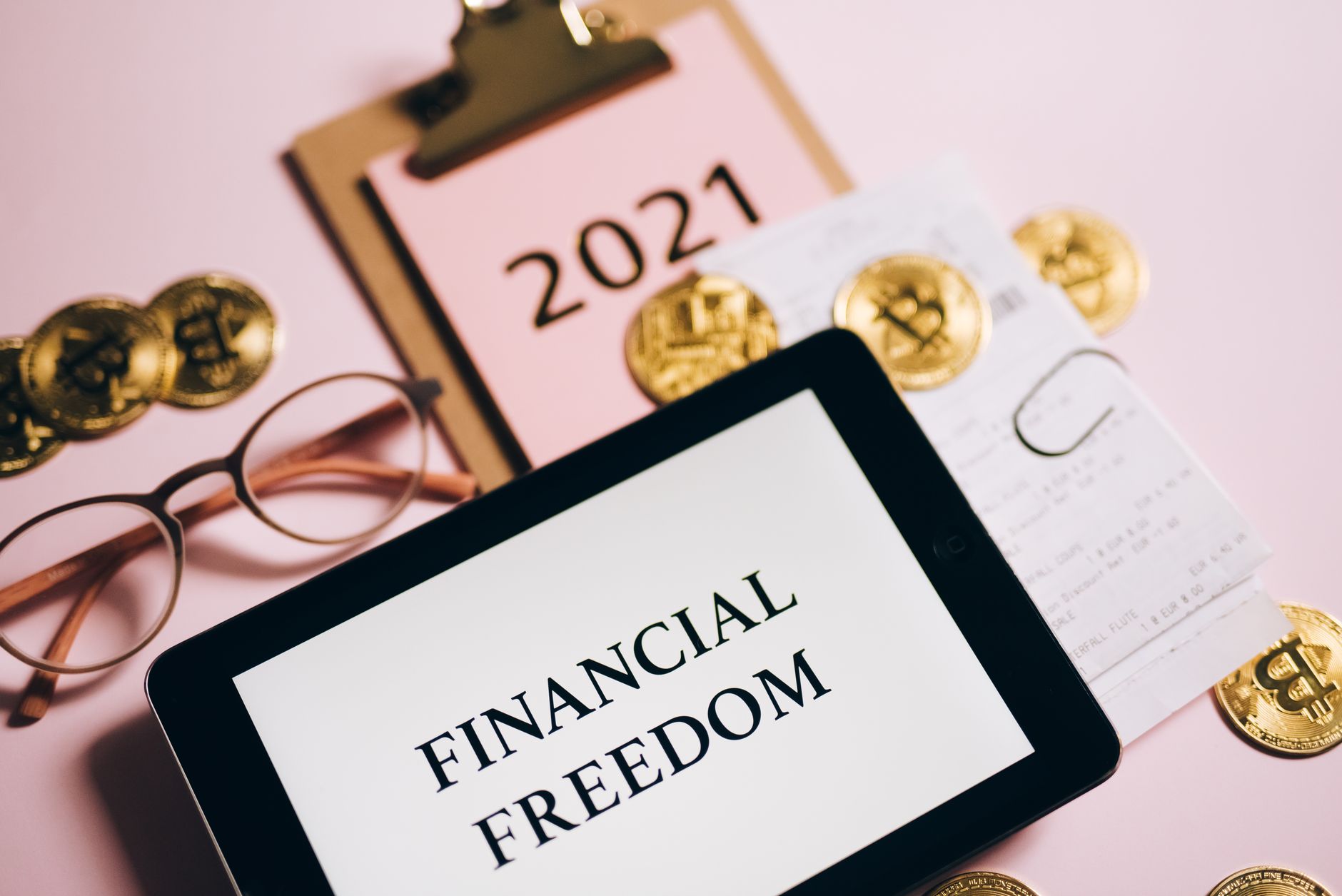 A tablet showing "Financial Freedom", a notebook, glasses, and gold bitcoin coins on a pink surface.