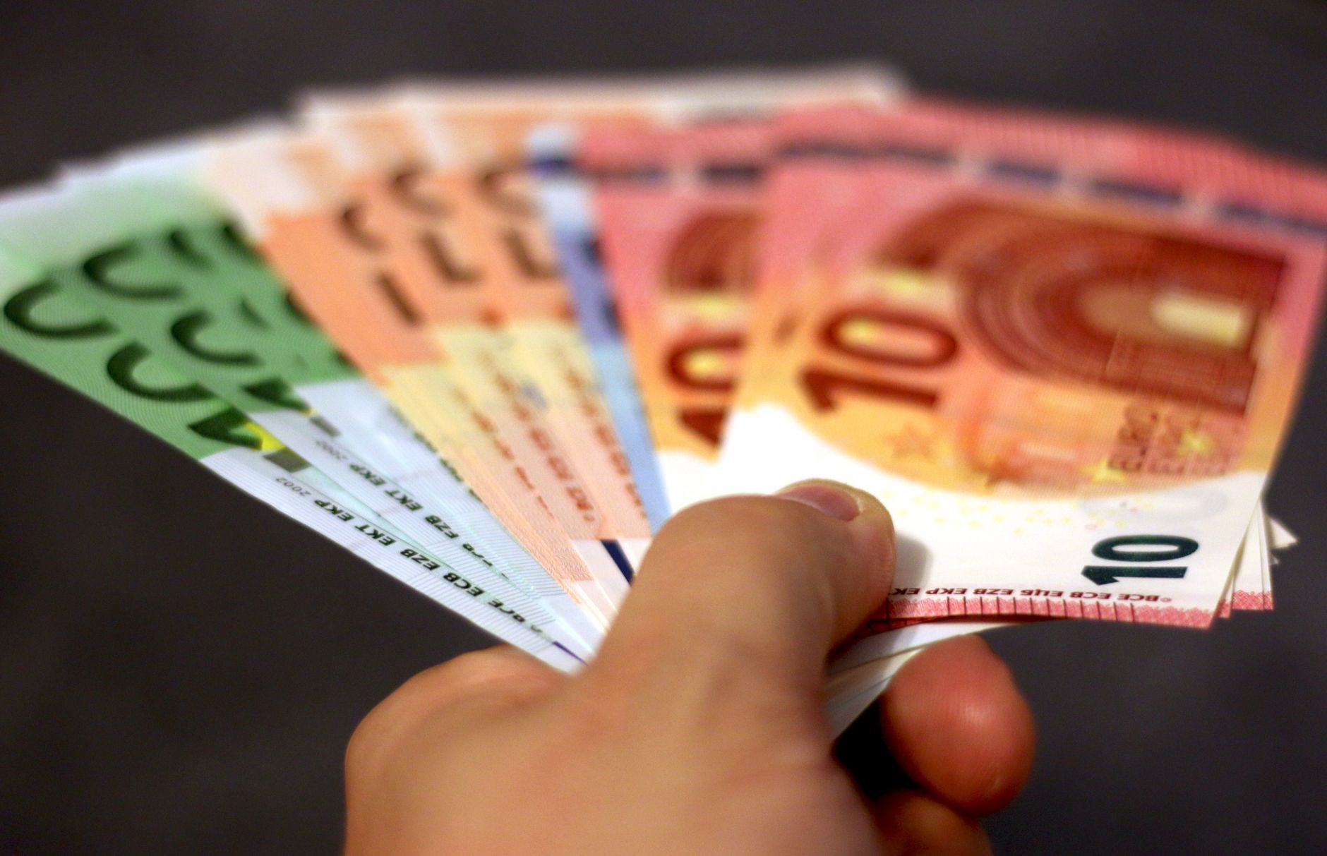 A hand holding a fan of Euro banknotes against a dark background for Cashflow Dana Pasif Bulanan.