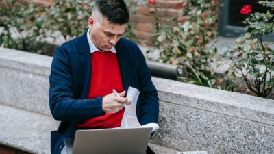 Seorang pria berjaket merah dan blazer biru duduk di bangku taman sambil bekerja dengan laptop dan dokumen.